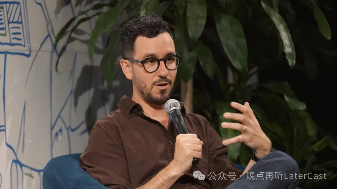 图片展示的是Anthropic的设计主管Joel Lewenstein。他身着棕色衬衫,戴着眼镜,坐在椅子上,左手持麦克风,右手比划着,似乎在讲解或讨论某个话题。背景为绿色植物和带有图案的白板。图片与上下文紧密相关,上下文提到Joel Lewenstein谈模型持续进步后,产品团队如何调整规划方式,此图可能是他在分享相关观点时的场景。
