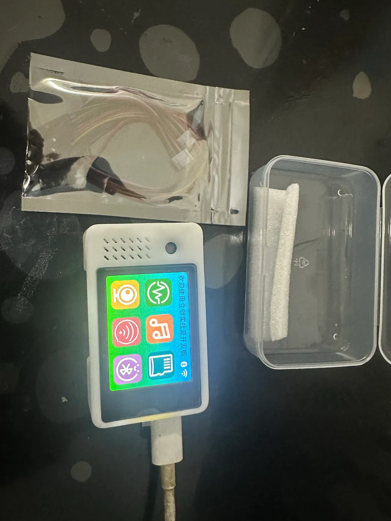 这张图片展示的是与ESP32开发相关的物品，主体是一台带彩色触控屏幕的一体开发板，屏幕上显示着六个彩色功能图标，开发板底部连接着一根白色的供电线。开发板旁放着一个密封袋，里面装有杜邦线，右侧还有一个透明收纳盒，盒内带有一块白色的缓冲垫。该图对应文档中介绍的N16R8一体开发板，呈现的是其开箱后的相关配件与设备本体，符合文档里提到的“开箱以后很简洁，一个小主机加几根杜邦线”的描述，也呼应了文档中提及的这款开发板适配相关项目且配套资源开源的内容。